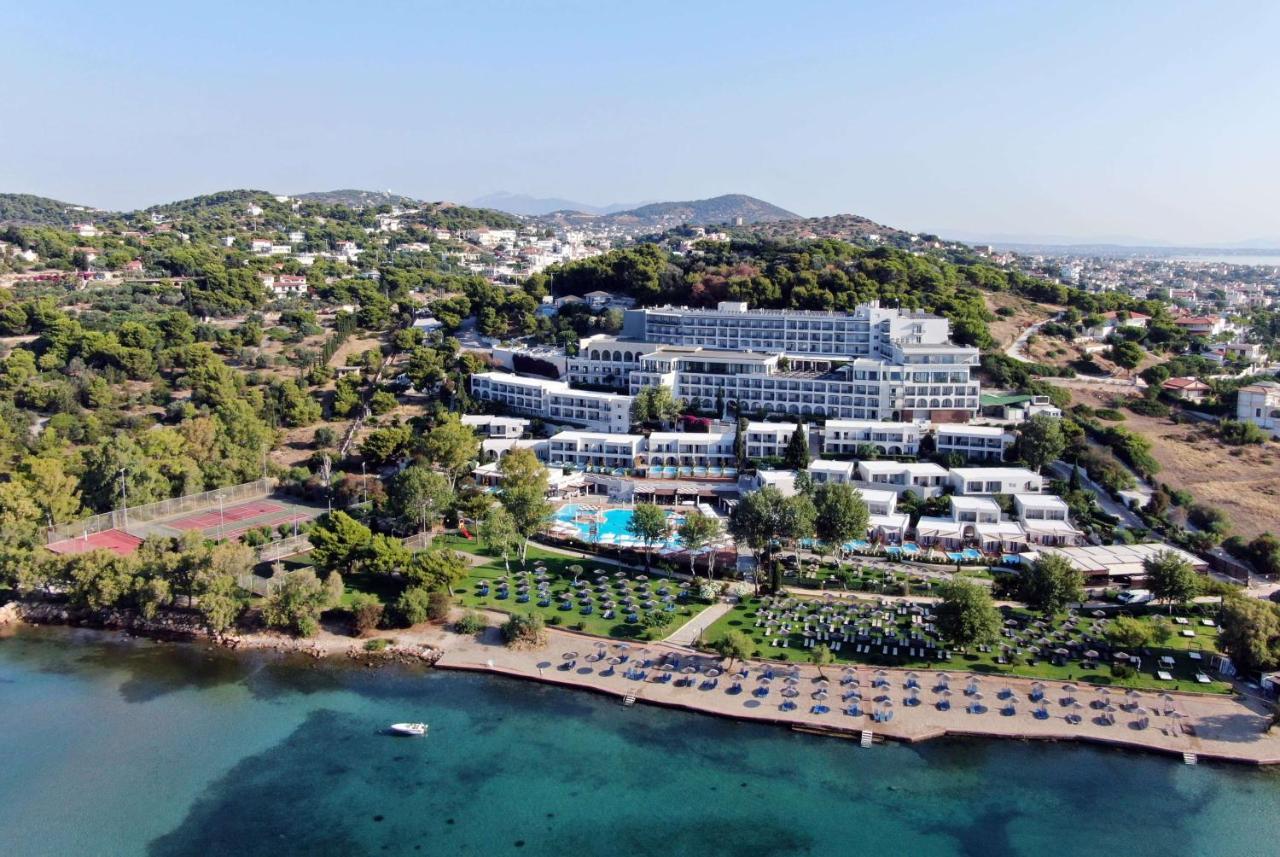 Dolce By Wyndham Athens Attica Riviera Hotel Vravrona Luaran gambar The image shows a large hotel complex situated near a beach. It's located on a hillside, surrounded by greenery and trees. The hotel features multiple levels with modern architecture, balconies, and a swimming pool area. In front of the hotel, there'