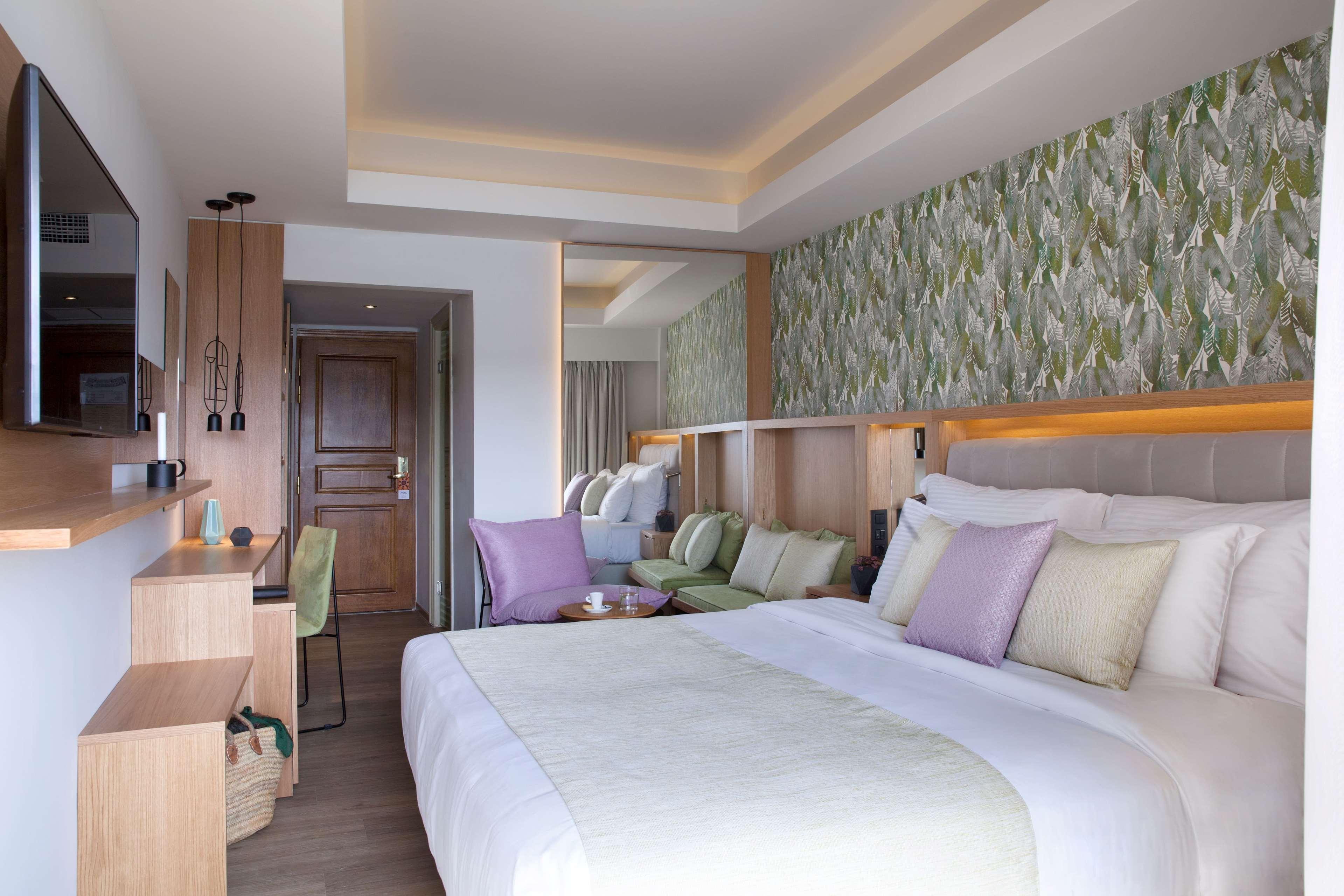 Dolce By Wyndham Athens Attica Riviera Hotel Vravrona Luaran gambar The photo shows a modern hotel room that features a spacious layout. There is a large bed with soft, neutral-colored linens and decorative pillows in shades of green and purple. Behind the bed, there is a stylish wallpaper with leafy patterns that ad