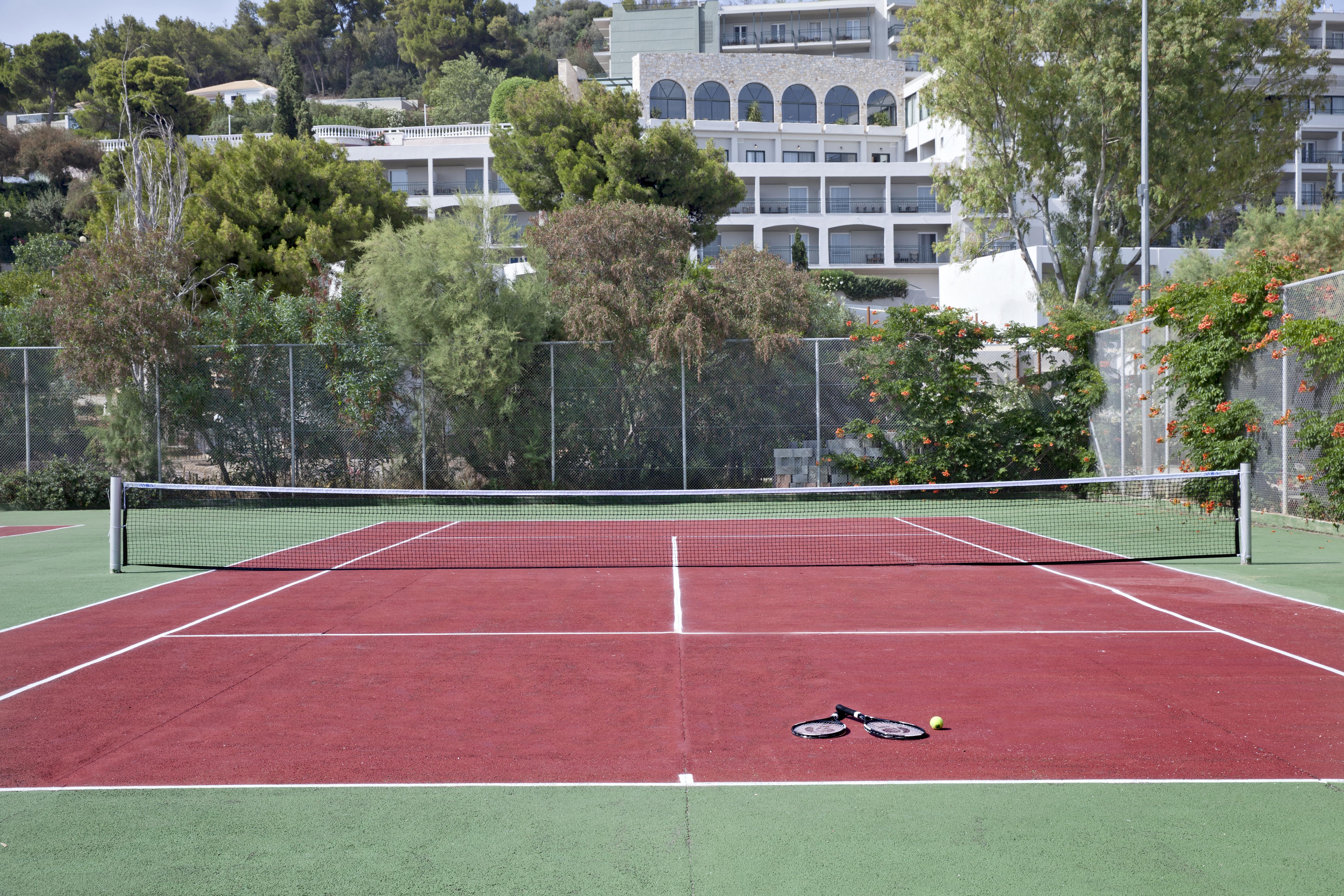 Dolce By Wyndham Athens Attica Riviera Hotel Vravrona Luaran gambar The photo shows a tennis court with red and green surfaces. The court is empty, with no players present. In the center, there are a tennis racket and a ball lying on the ground. In the background, there are trees and a building, suggesting the court 