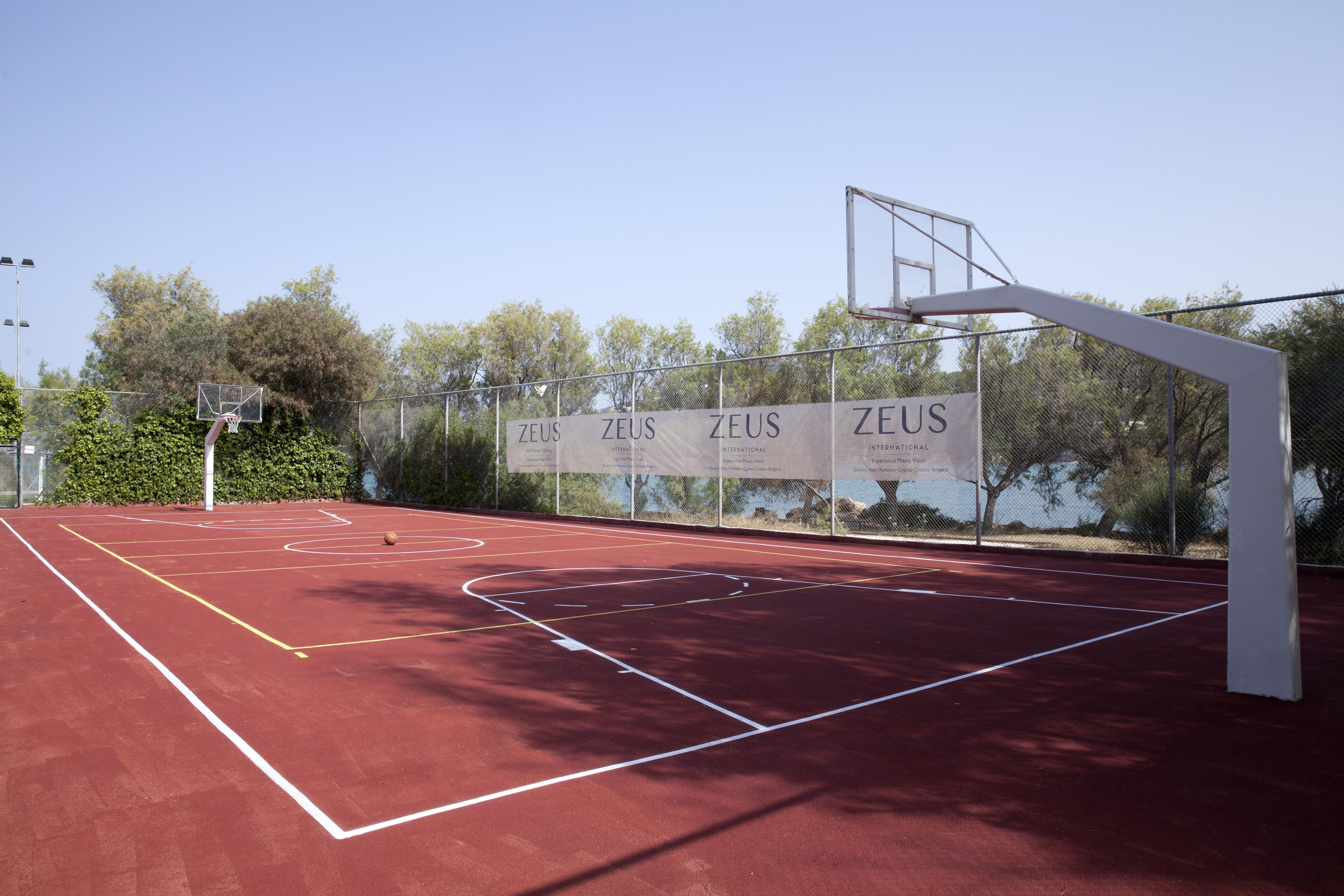 Dolce By Wyndham Athens Attica Riviera Hotel Vravrona Luaran gambar The photo shows a basketball court with a reddish surface. There are two basketball hoops on either side of the court, surrounded by a fence that has banners displaying the word "ZEUS." In the background, there are trees, and the court appears to be 