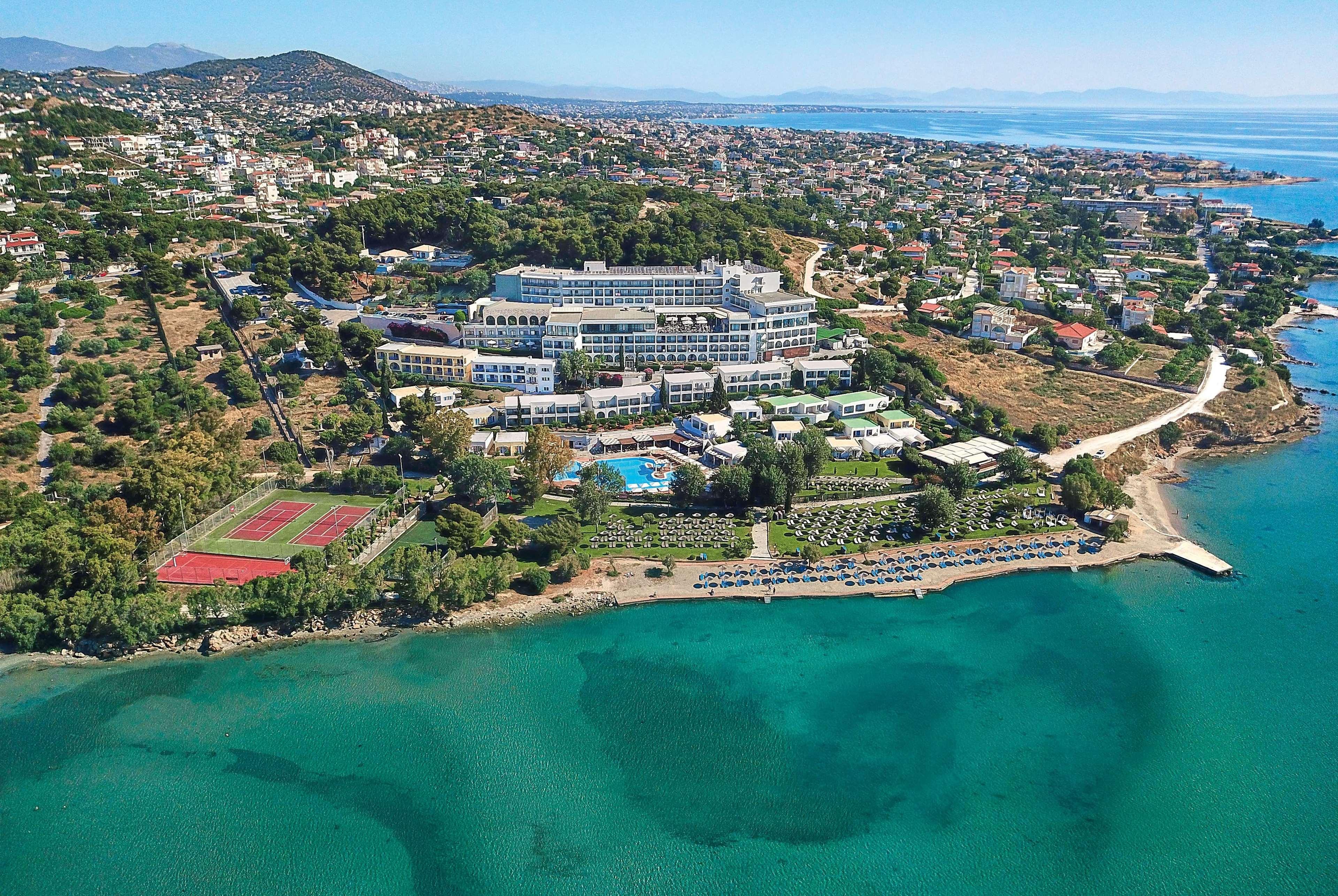 Dolce By Wyndham Athens Attica Riviera Hotel Vravrona Luaran gambar The photo shows an aerial view of a coastal resort area. The resort features multiple buildings, including hotel accommodations and amenities, situated on a hillside overlooking a calm, turquoise sea. There is a sandy shoreline with a few boats on th
