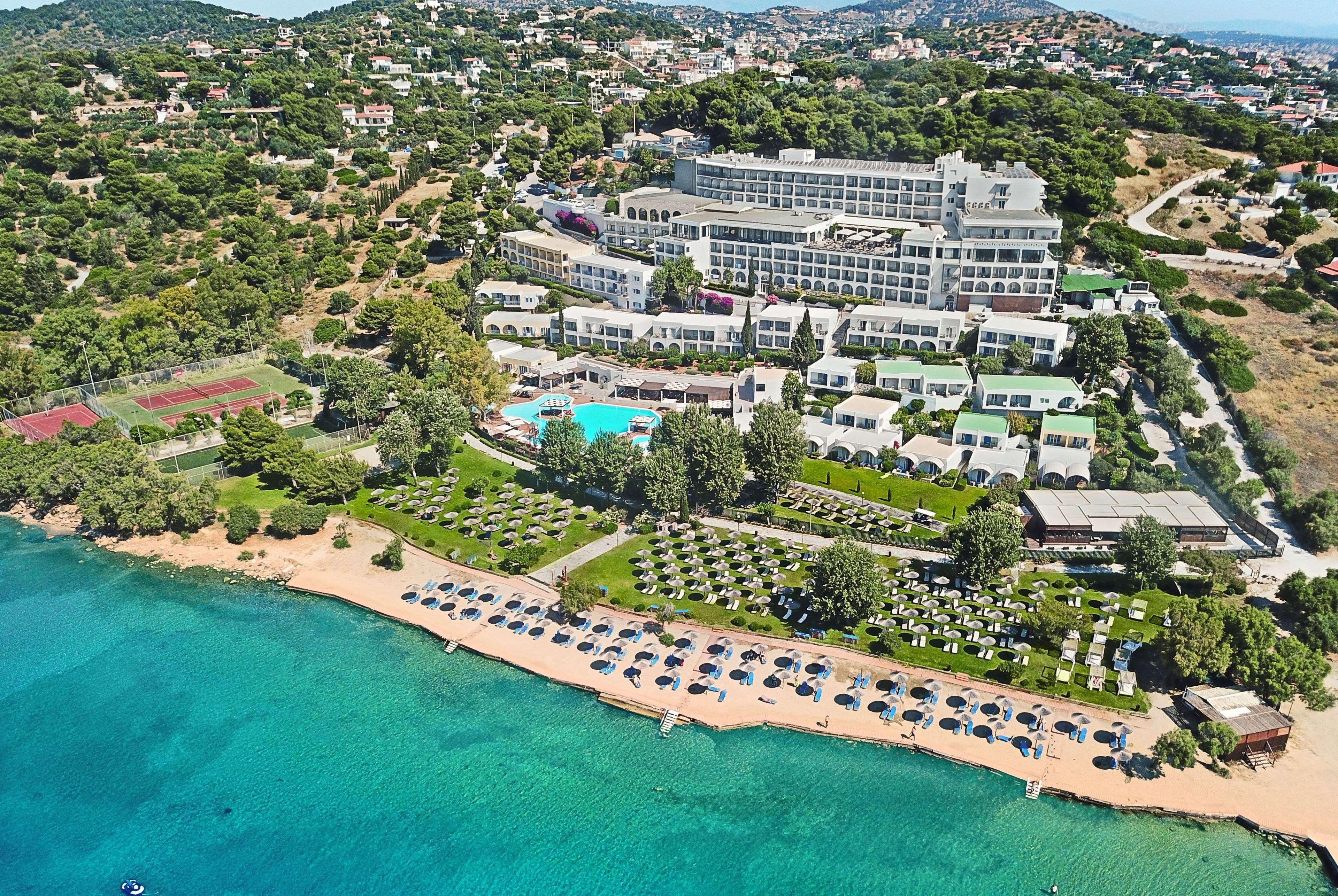 Dolce By Wyndham Athens Attica Riviera Hotel Vravrona Luaran gambar The photo shows a scenic aerial view of a resort located near a beautiful coastline. The resort features a large building and various smaller accommodations. An inviting swimming pool is visible at the center, surrounded by sun loungers and greenery.