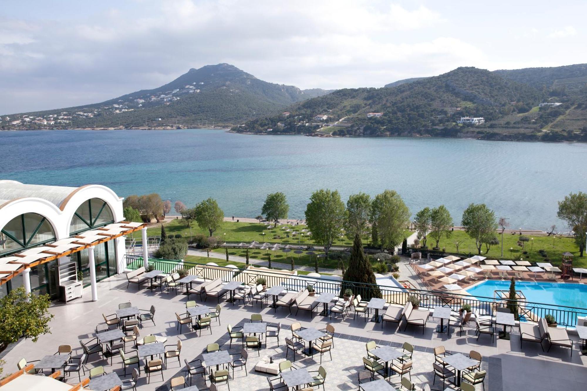 Dolce By Wyndham Athens Attica Riviera Hotel Vravrona Luaran gambar The photo depicts a scenic view from a terrace or outdoor dining area. In the foreground, there are numerous tables and chairs arranged neatly, likely for guests to enjoy meals or drinks. Beyond the seating area, a swimming pool can be seen. 

In the