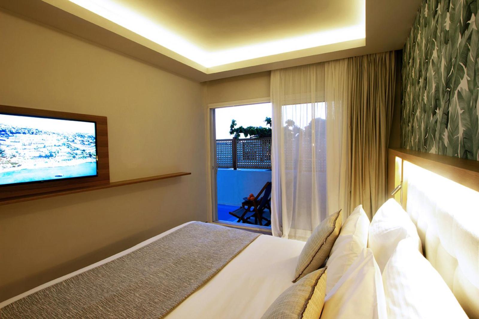 Dolce By Wyndham Athens Attica Riviera Hotel Vravrona Luaran gambar The photo shows a modern hotel room featuring a large bed with white linens and decorative pillows. There's a mounted flat-screen television on the wall opposite the bed. Natural light enters the room through sheer curtains, leading to a balcony or o